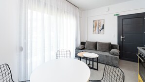 A modern living room with a sofa, a dining table, and chairs. There is a painting on the wall and a door with an exit sign. - White Dream One-bedroom Apartment With Pool Access (Thasos)