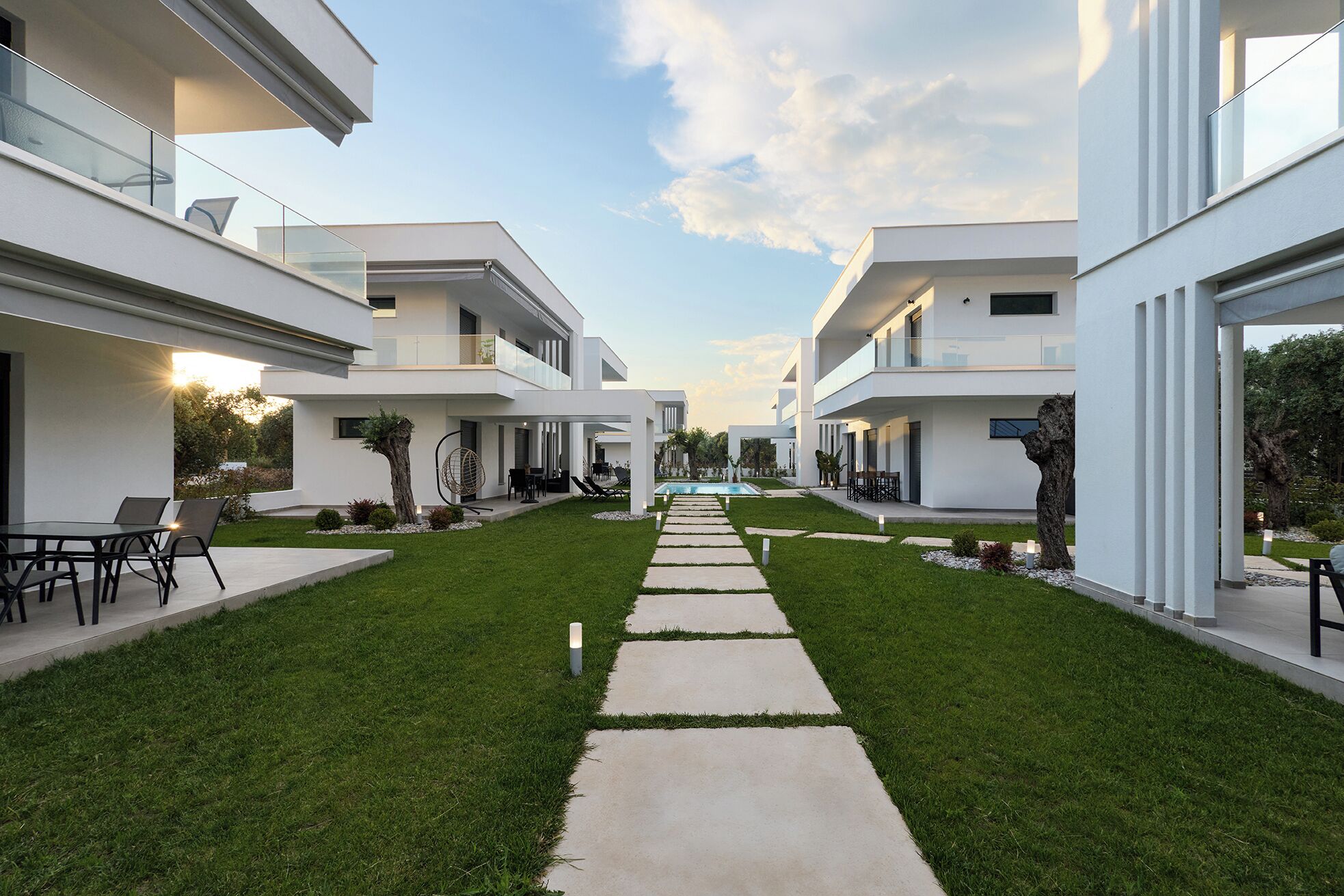 A modern residential complex with a central pathway leading to a swimming pool, surrounded by well-maintained lawns and outdoor seating areas.