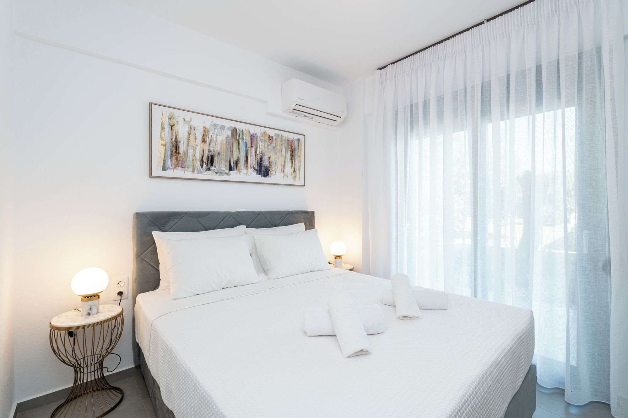 Une chambre d’hôtel avec un grand lit, du linge de lit blanc, un tableau abstrait encadré, un appareil de climatisation et des rideaux en voile.