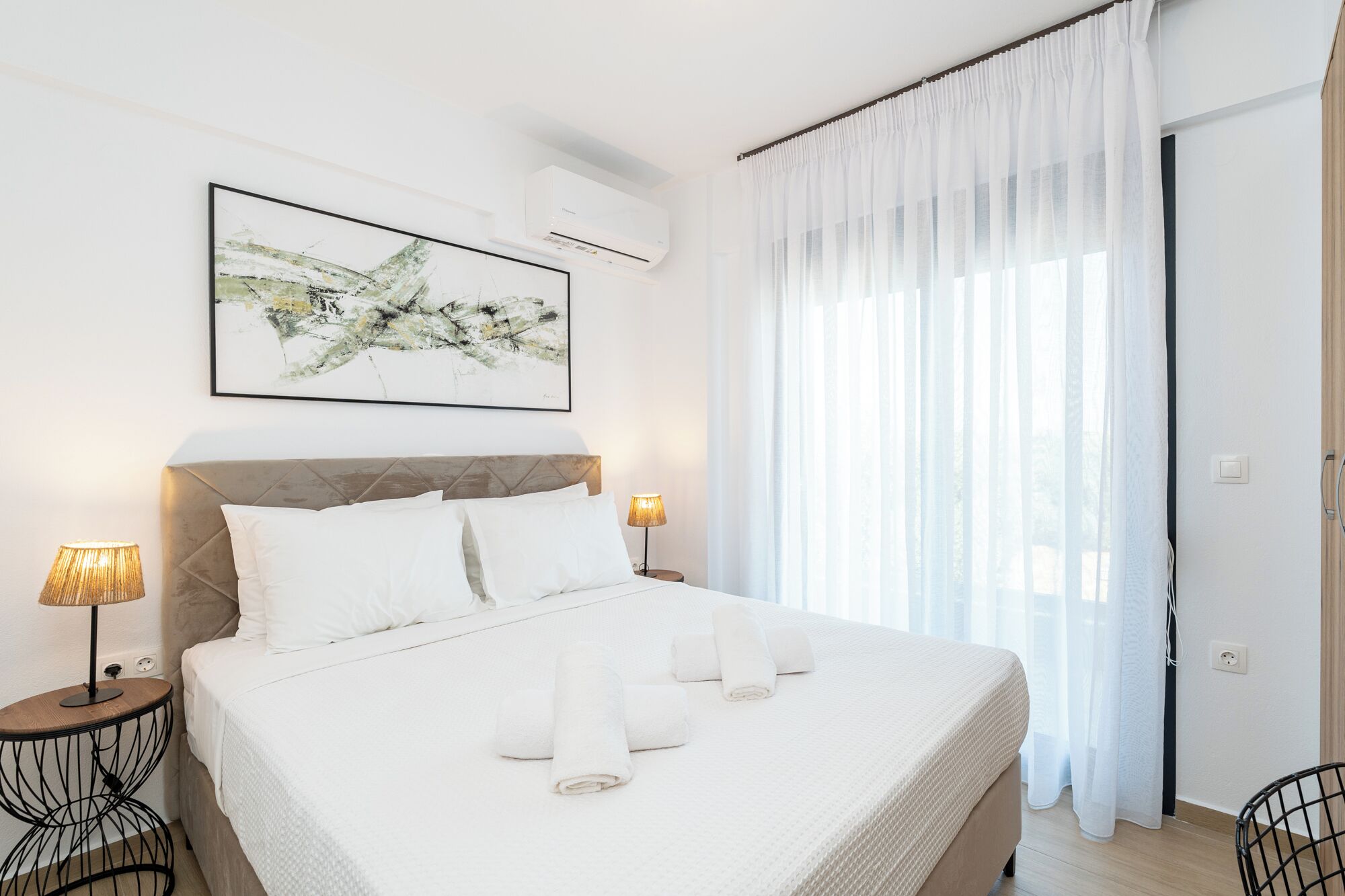 A neatly made bed with white linens and rolled towels, a modern headboard, a wall-mounted air conditioner, and a large window with sheer curtains.