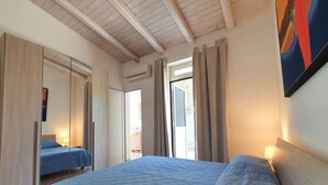 Et soveværelse med en stor seng, et garderobeskab, en dør der fører til en balkon og et loft med træbjælker.