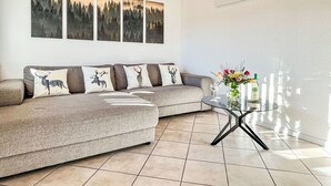 Una sala moderna con un sofá gris, cuatro cuadros con motivos forestales colgados en la pared, una mesa de centro de cristal con copas y botellas de vino, e iluminación colgante.
