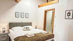 Un dormitorio con cama, mesitas de noche, una viga de madera en el techo, cuadros enmarcados en la pared y una puerta que da al baño.
