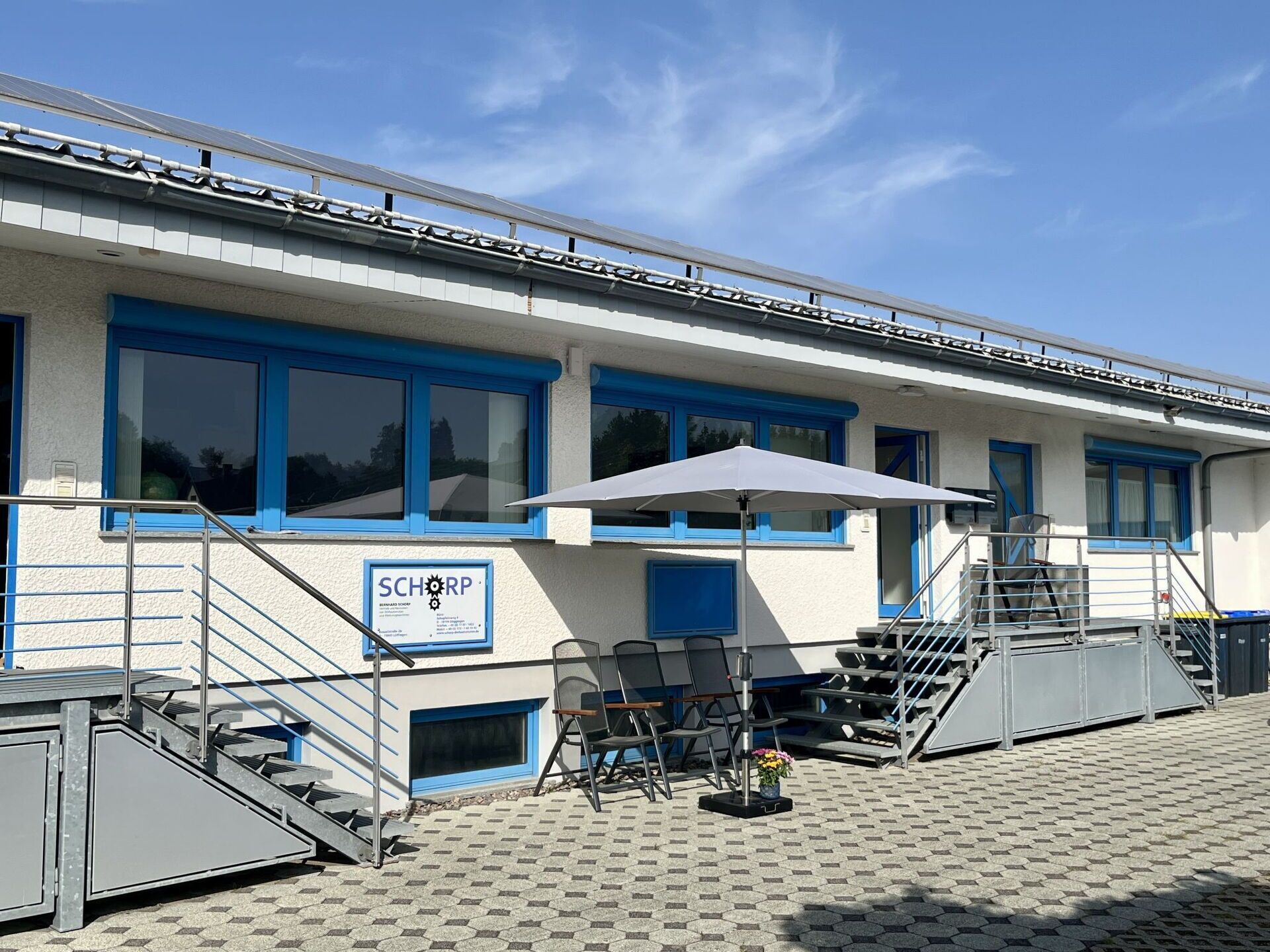 A building with blue-framed windows, a signboard, outdoor seating with chairs and a table, and a staircase leading to an upper level.
