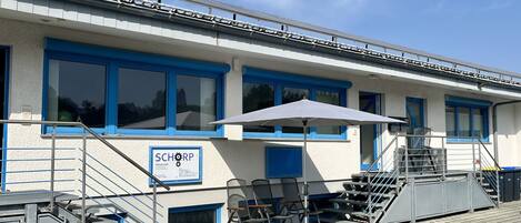 A building with blue-framed windows, a signboard, outdoor seating with chairs and a table, and a staircase leading to an upper level.