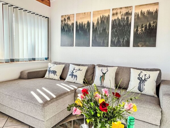 Una sala de estar con un sofá gris, tres cojines con estampados de ciervos, un jarrón de flores y cuatro cuadros con motivos forestales en la pared.