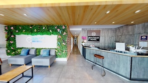 Un espace d’accueil d’hôtel au design moderne, comprenant un comptoir d’accueil, un coin salon et un mur agrémenté de plantes et de fleurs.