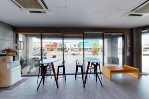 Un moderno vestíbulo de hotel con un separador de vidrio, una mesa de recepción y un área de descanso con una mesa redonda y sillas.