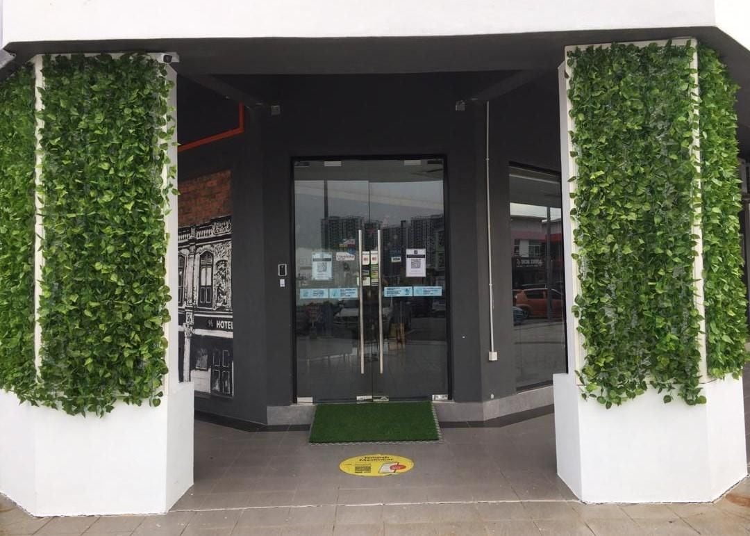 A hotel entrance with a glass door, a green mat at the entrance, and a wall with greenery on either side.