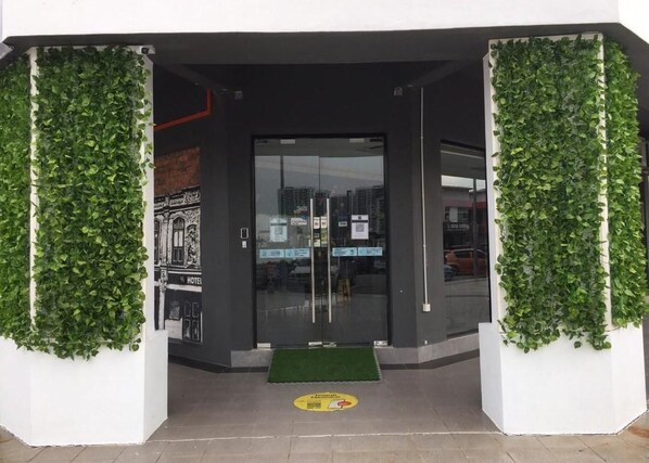 A hotel entrance with a glass door, a green mat at the entrance, and a wall with greenery on either side.