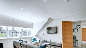 A modern living room with a grey sofa, a TV mounted on the wall, and a large window offering a city view.