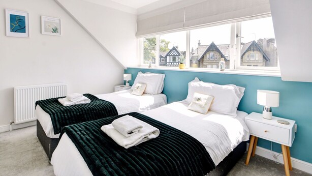 A bedroom with two beds, a window with a view of houses, a radiator, and a bedside table with a lamp.