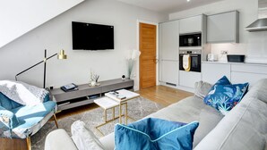 A modern living room with a sofa, coffee table, and a TV. A kitchen is visible with appliances and a microwave.