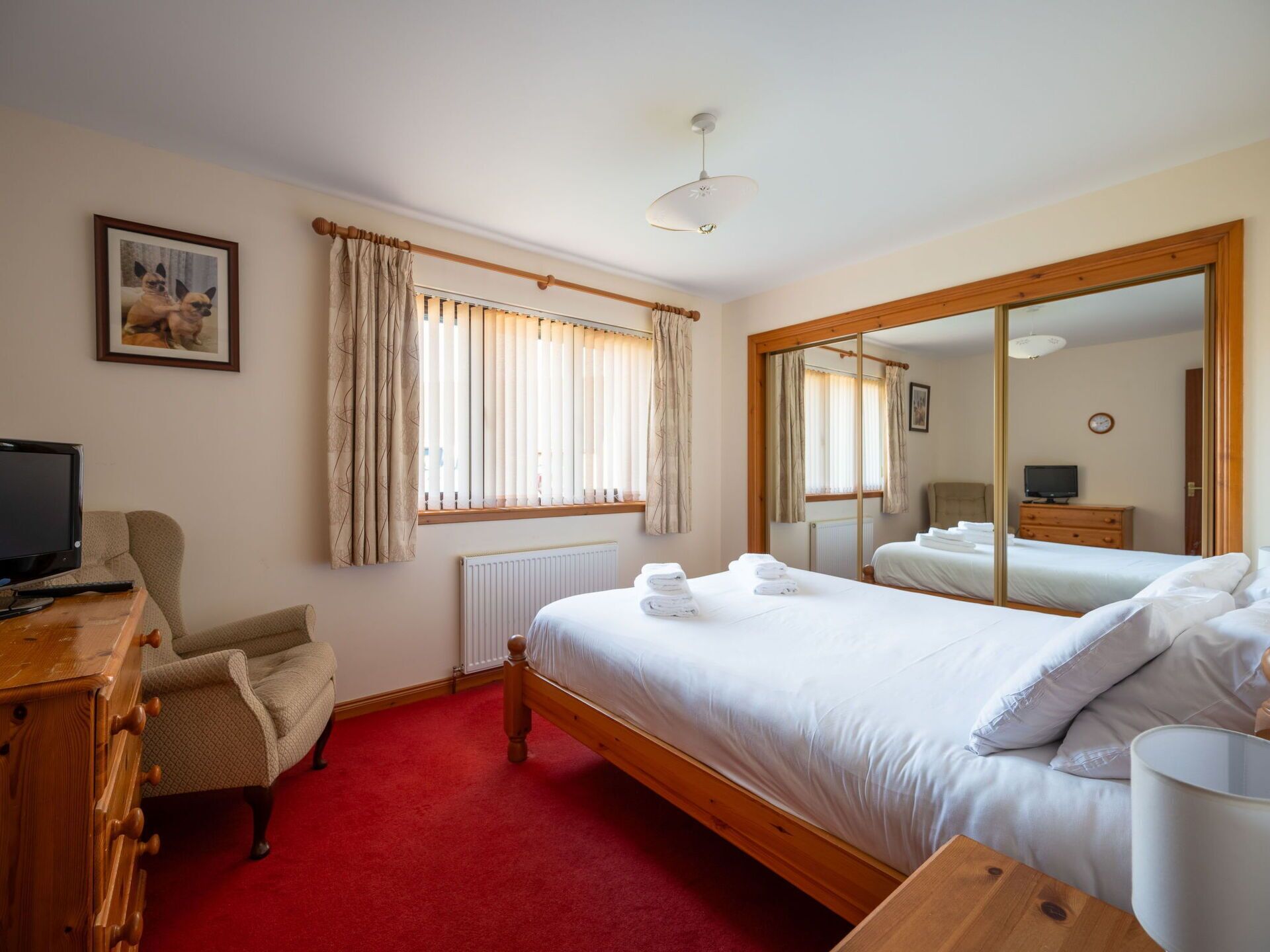 A hotel room with a bed, a desk with a computer, a chair, a large mirror, and a window with curtains.