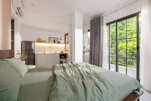 Une chambre avec un lit, une vue sur la verdure par la fenêtre, un coin cuisine et une salle de bain.