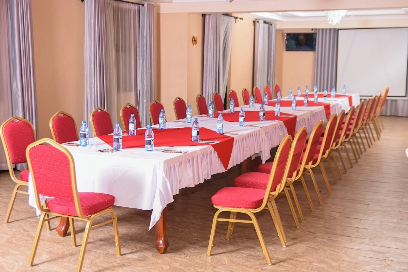 A conference room with rows of tables and chairs, water bottles on tables, and a projector screen in the background.