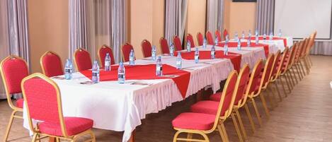 A conference room with rows of tables and chairs, water bottles on tables, and a projector screen in the background.