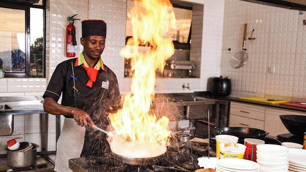 A chef cooking with flames, a fire extinguisher, and kitchen utensils in a commercial kitchen. - Golden Palace Hotel Eldoret (Eldoret)