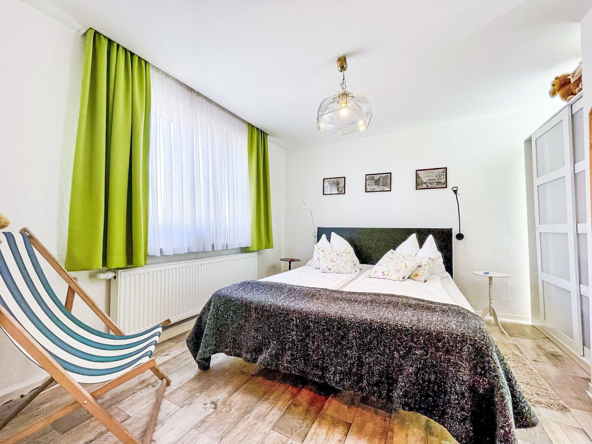 Een slaapkamer met een bed, een stoel, een raam met gordijnen, een wandlamp en drie foto's.
