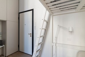Une pièce avec une porte blanche, une échelle blanche, un bureau blanc et un plafond blanc percé d’une grille d’aération.