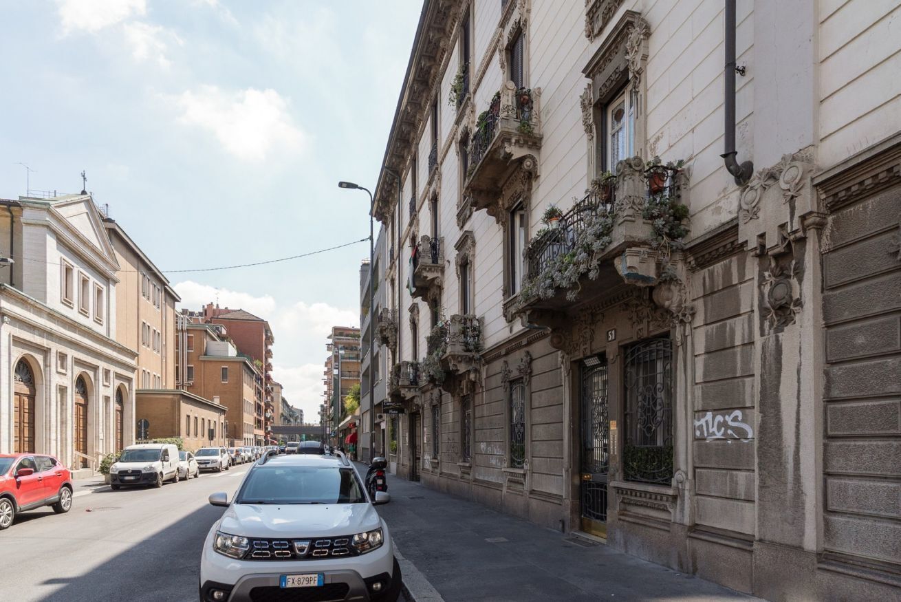 Une vue de rue avec une voiture blanche garée sur le bord, des bâtiments aux façades richement décorées et un ciel dégagé.