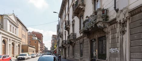 A street view with a white car parked on the side, buildings with ornate facades, and a clear sky.