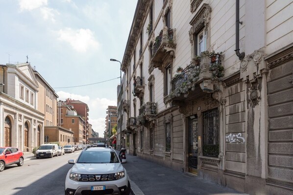 A street view with a white car parked on the side, buildings with ornate facades, and a clear sky.