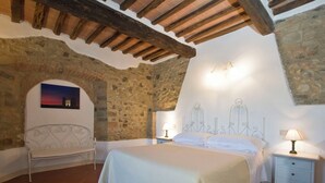 A bedroom with a stone wall, a bed with a white bedspread, a bench, and a nightstand with a lamp. - Cosy apartment in villa for 6 guests with WIFI, pool, TV, terrace, pets allowed and panoramic view (San Giovanni D'asso)