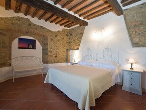 A bedroom with a stone wall, a bed with a white bedspread, a bench, and a nightstand with a lamp.