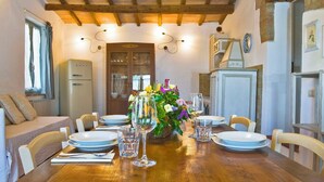 Un'area pranzo con un tavolo di legno apparecchiato per quattro persone, circondato da sedie bianche. Si notano, sullo sfondo, un frigorifero e un armadietto.