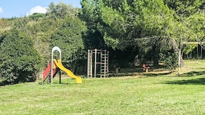Ein Spielplatz mit Rutsche und Klettergerüsten in einer grünen Parklandschaft mit Bäumen.