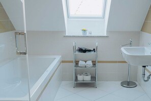 Ein modernes Badezimmer mit einer Badewanne, einem Waschbecken, einem Ablagefach mit Handtüchern und Pflegeprodukten sowie einem Fenster über dem Waschbecken.