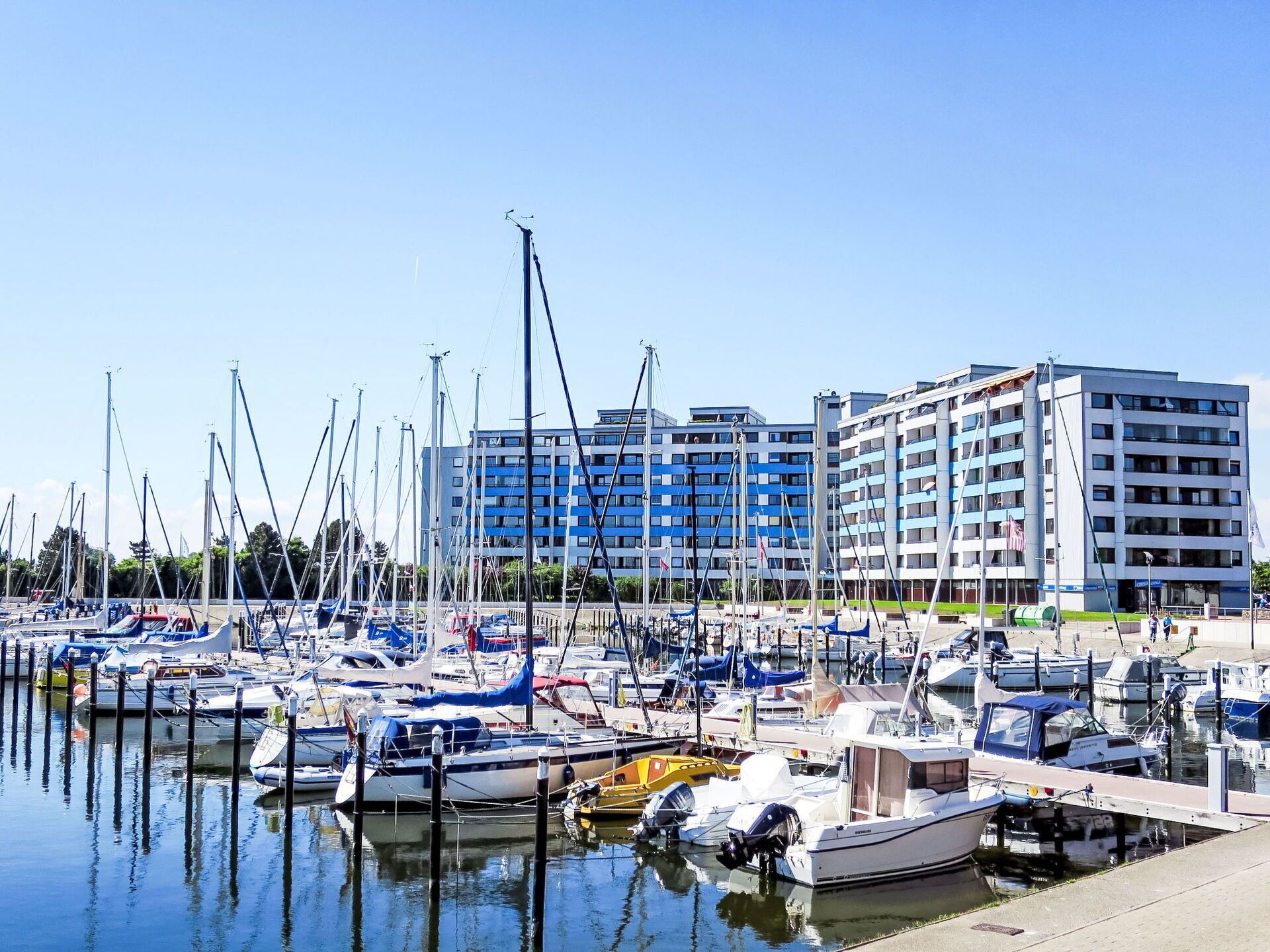 Un port de plaisance où sont amarrés différents types de bateaux, un grand immeuble à étages avec de nombreux balcons, et un ciel bleu dégagé.
