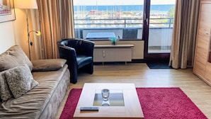 Une chambre d’hôtel équipée d’un canapé, d’un fauteuil, d’une table basse avec une télécommande, d’une fenêtre donnant sur un port de plaisance et d’un radiateur.
