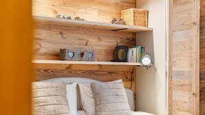 Een slaapkamer met een houten hoofdbord, een bed met beige beddengoed en een plank boven het bed met boeken en decoratieve objecten.