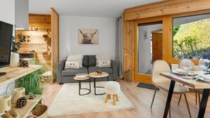 Een gezellige woonkamer met een grijze bank, een klein rond tafeltje en een houten eettafel met stoelen. Links staat een televisie, een houten trap en een groot raam met uitzicht op bomen.