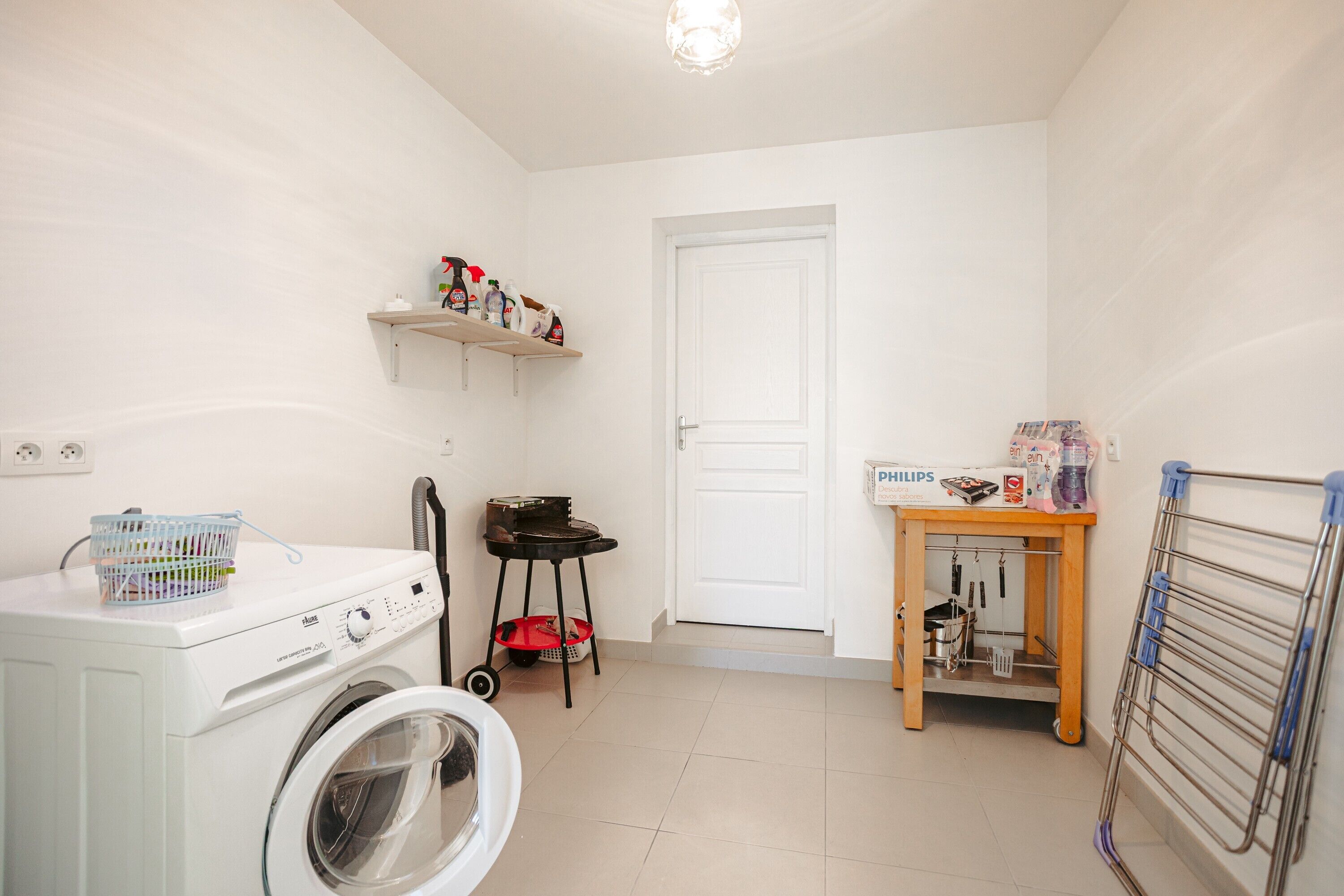 A laundry area with a washing machine, a drying rack, a small table with cleaning supplies, and a shelf with detergent bottles.