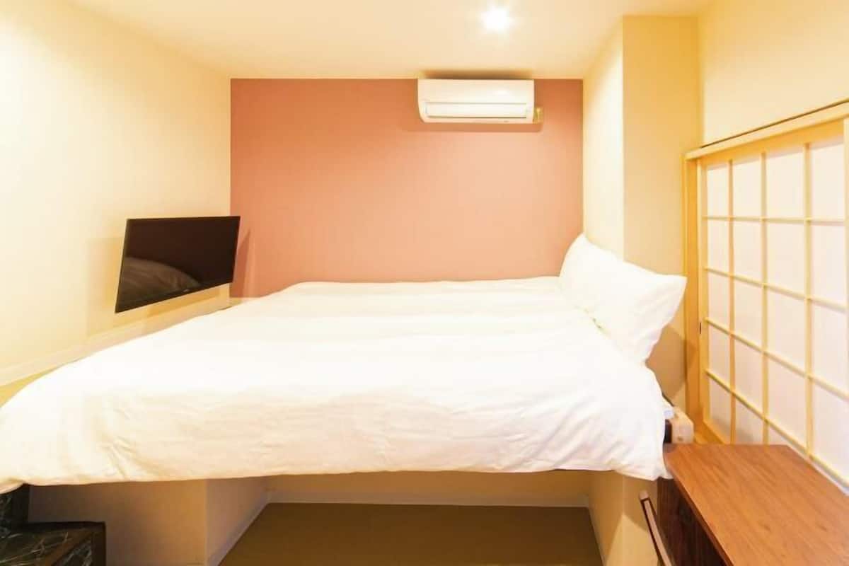 Una cama individual con colchón y almohadas blancos, un cabecero de madera, un aire acondicionado instalado en la pared y una pequeña estantería de madera.