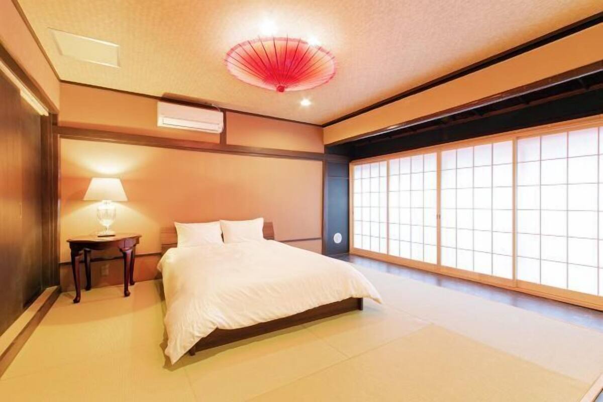 Un dormitorio japonés tradicional con suelo de tatami, una cama con ropa de cama blanca, una mesita de noche con lámpara y una puerta corredera de shoji.