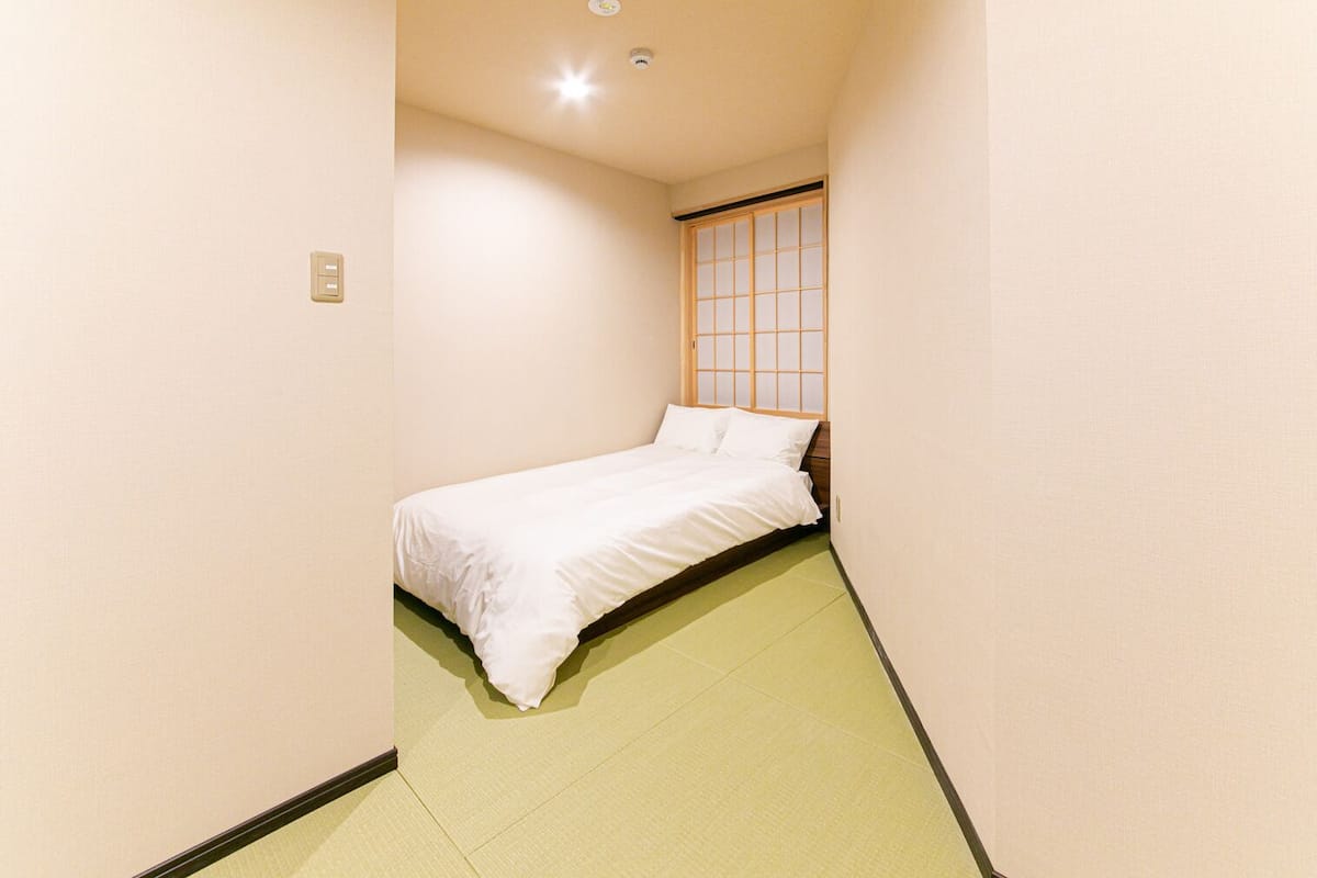 Una habitación estrecha con una cama, una ventana de diseño de cuadrícula y un interruptor de luz en la pared.