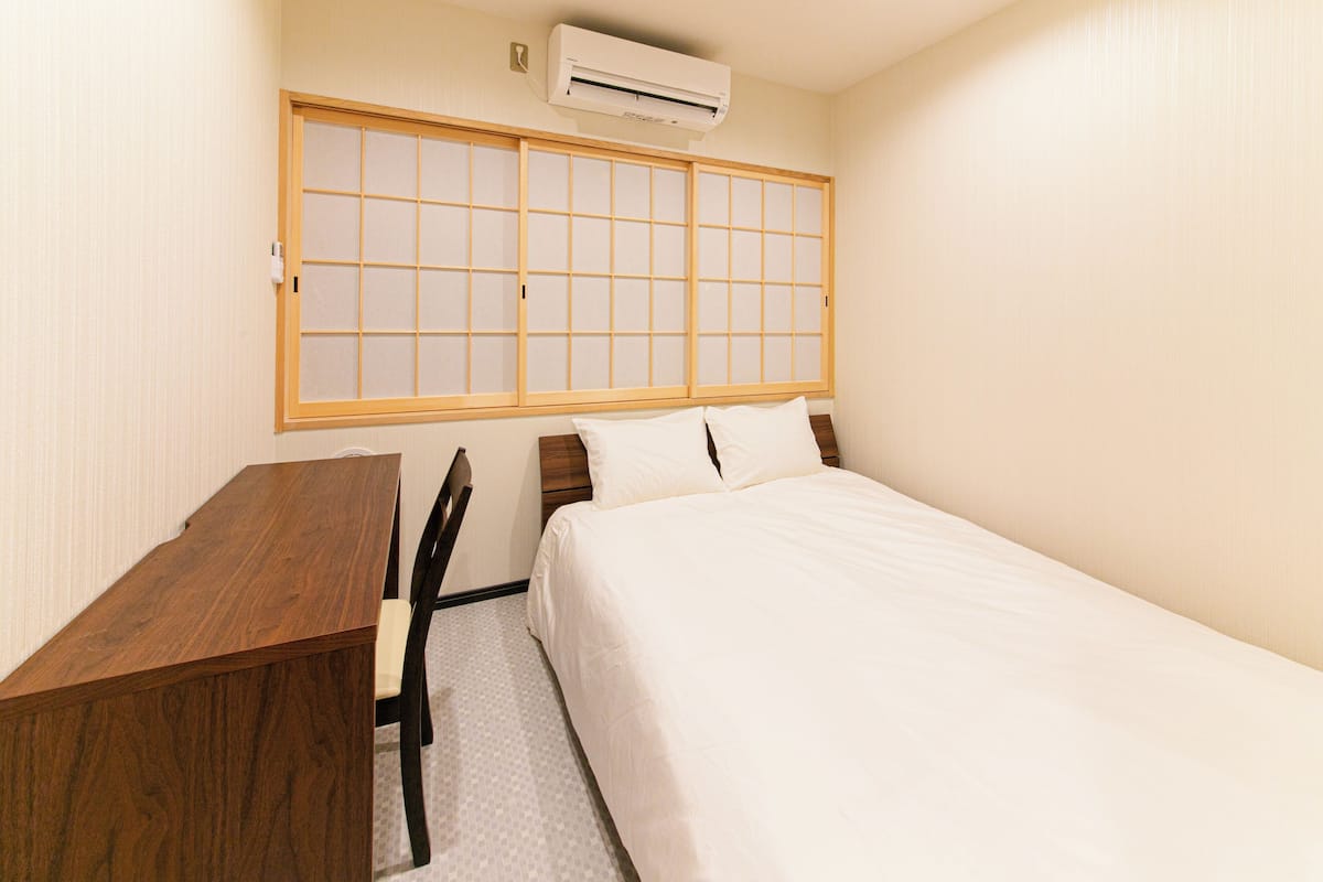 Una habitación de hotel con un escritorio de madera, una cama con ropa blanca y un ventanal con diseño de cuadrícula.