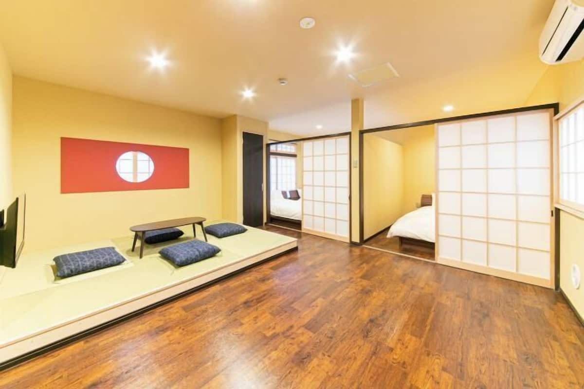 Una habitación con zona de estar en tatami, una cama con celosía de papel y una bandera roja en la pared.