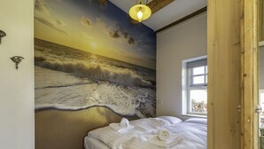 A bedroom with a mural of a beach scene, a bed with white linens, and a window with a view of greenery outside.