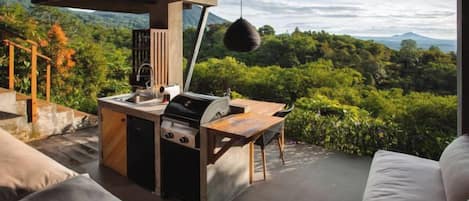 An outdoor kitchen with a grill, sink, and hanging light fixture, overlooking a lush landscape.