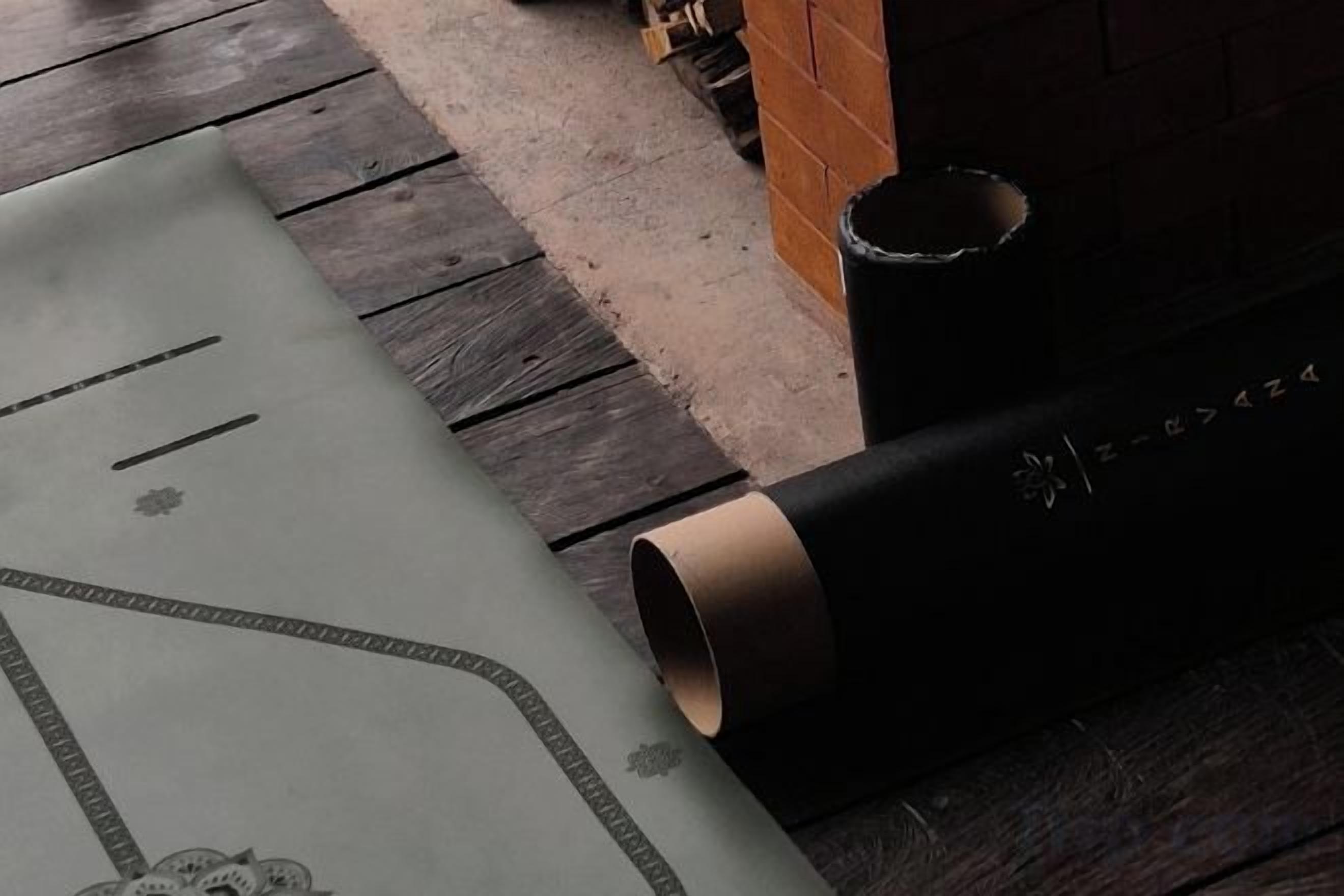 Un tapete enrollado con un diseño geométrico, un objeto cilíndrico con superficie texturizada y una pared de ladrillo al fondo.