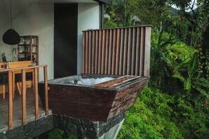 A spa area with a wooden tub on a stone platform, surrounded by lush greenery. - Aurora Cabins by Panoramika (Bali)