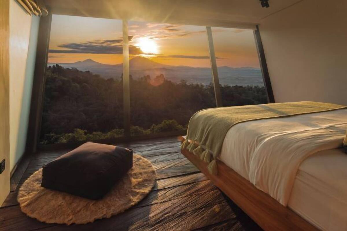 Een slaapkamer met een groot raam met uitzicht op een bergachtig landschap bij zonsondergang.