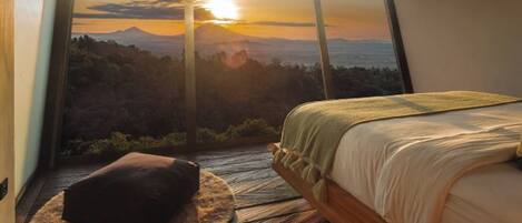 A bedroom with a large window overlooking a mountainous landscape at sunset.