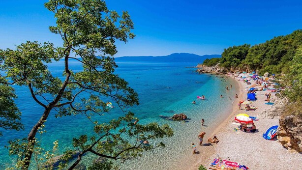 Ein Strand mit klarem Wasser, badenden Personen und Sonnenschirmen.