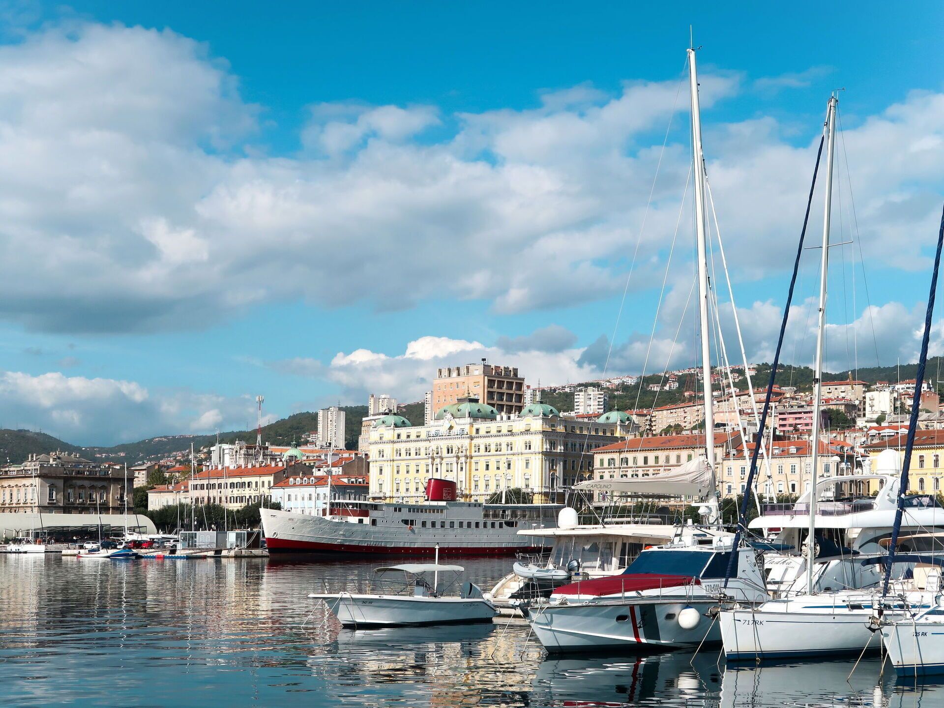 A marina with several boats docked, a large cruise ship, and buildings in the background.
